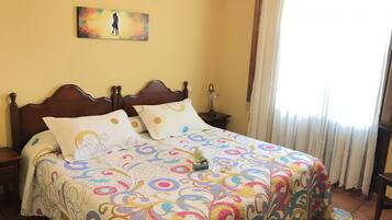 A bedroom with a wooden bed, colorful bedding, a nightstand with a lamp, a painting on the wall, and a window with curtains.