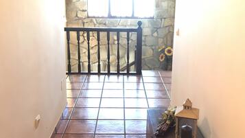 Un pasillo con piso de baldosas, pared de piedra, barandal negro y un farol sobre una mesa.