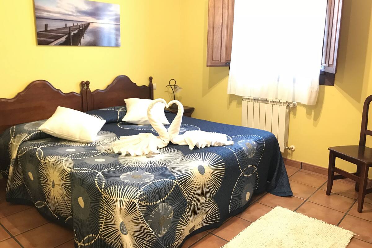 Ein Schlafzimmer mit einem Bett, einem Holz-Kopfteil, einem Wandbild, einem Fenster mit Vorhängen und einem Stuhl.