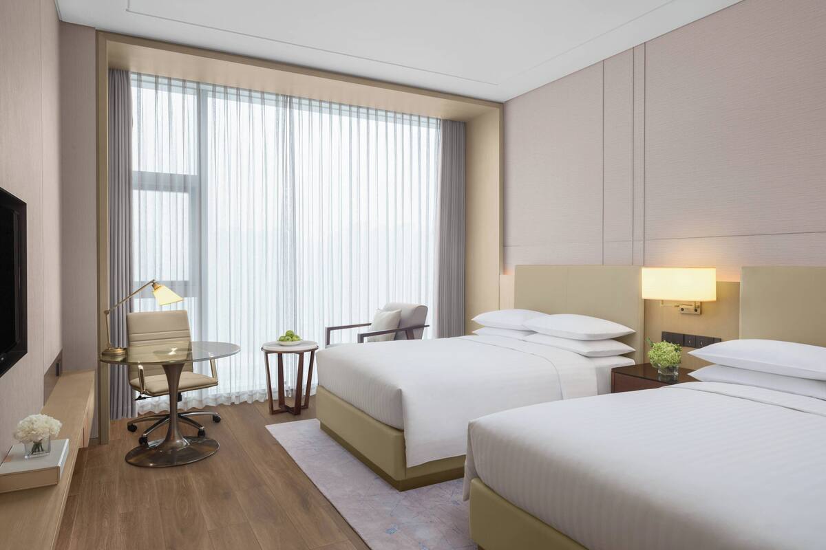 A hotel room with two beds, a desk with a chair, a TV, and a window with curtains.