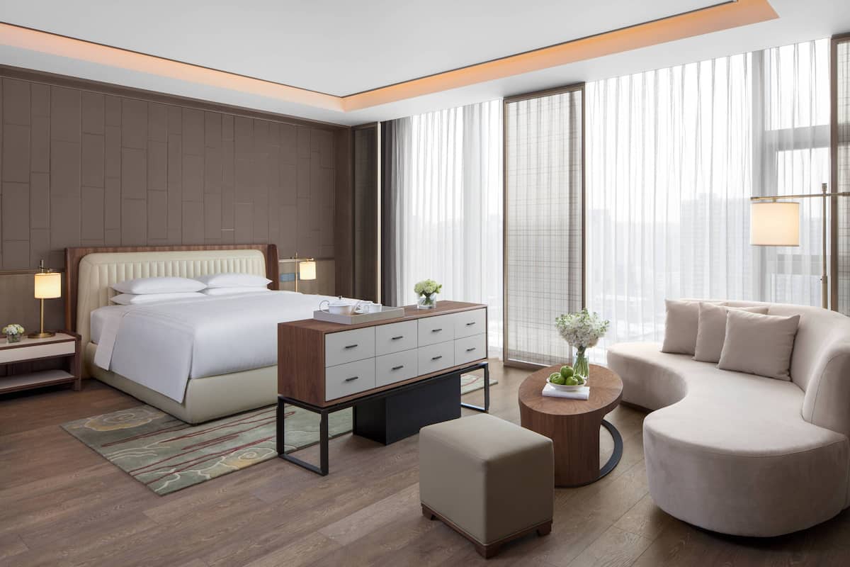 A modern hotel room with a large bed, a sofa, a small table, and a nightstand with a lamp.