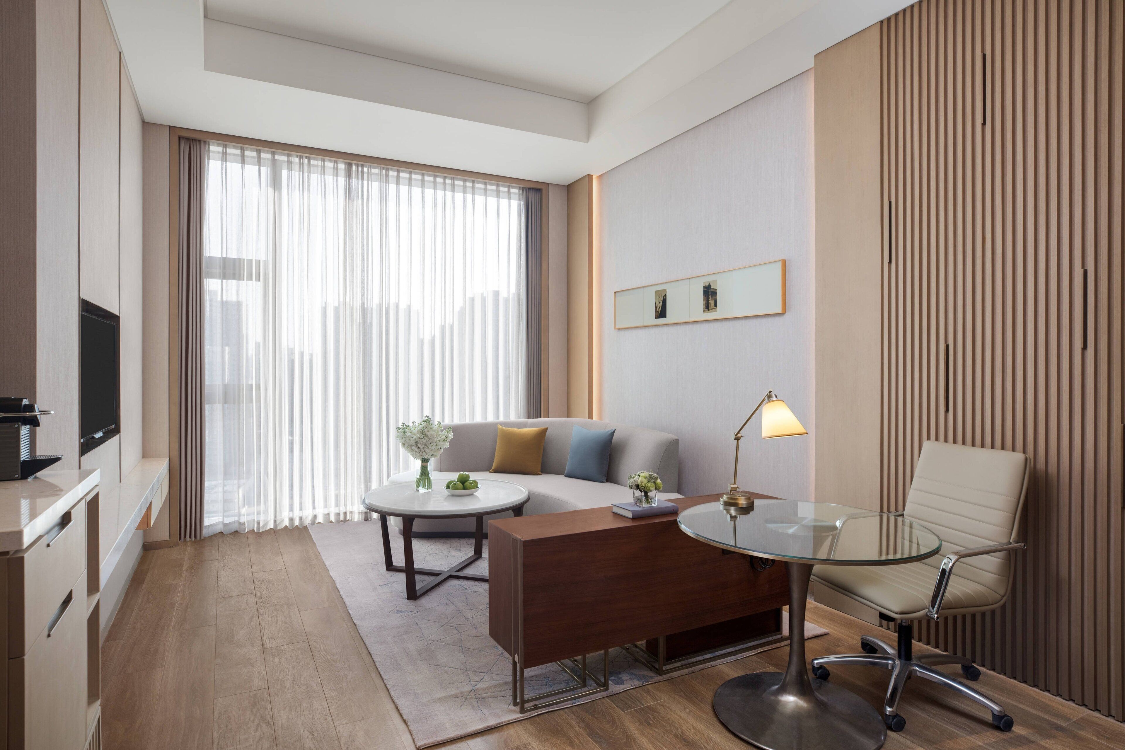 A modern hotel room with a sofa, a glass table, a chair, a TV, and a large window with curtains.