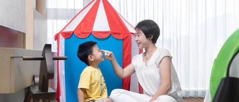 A room with a child and an adult sitting on the floor, a small tent-like structure, and a wooden chair.
