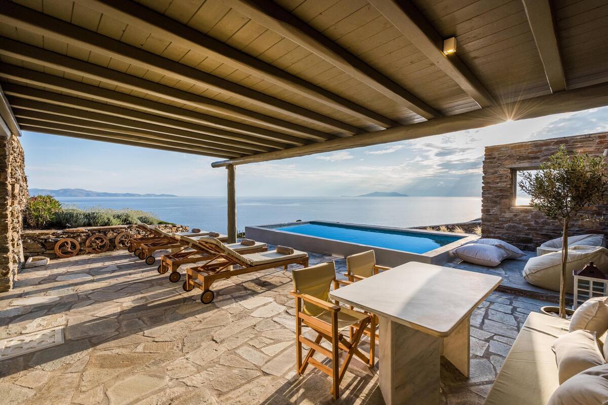 An outdoor patio with a stone floor, wooden furniture, a pool, and a view of the sea.