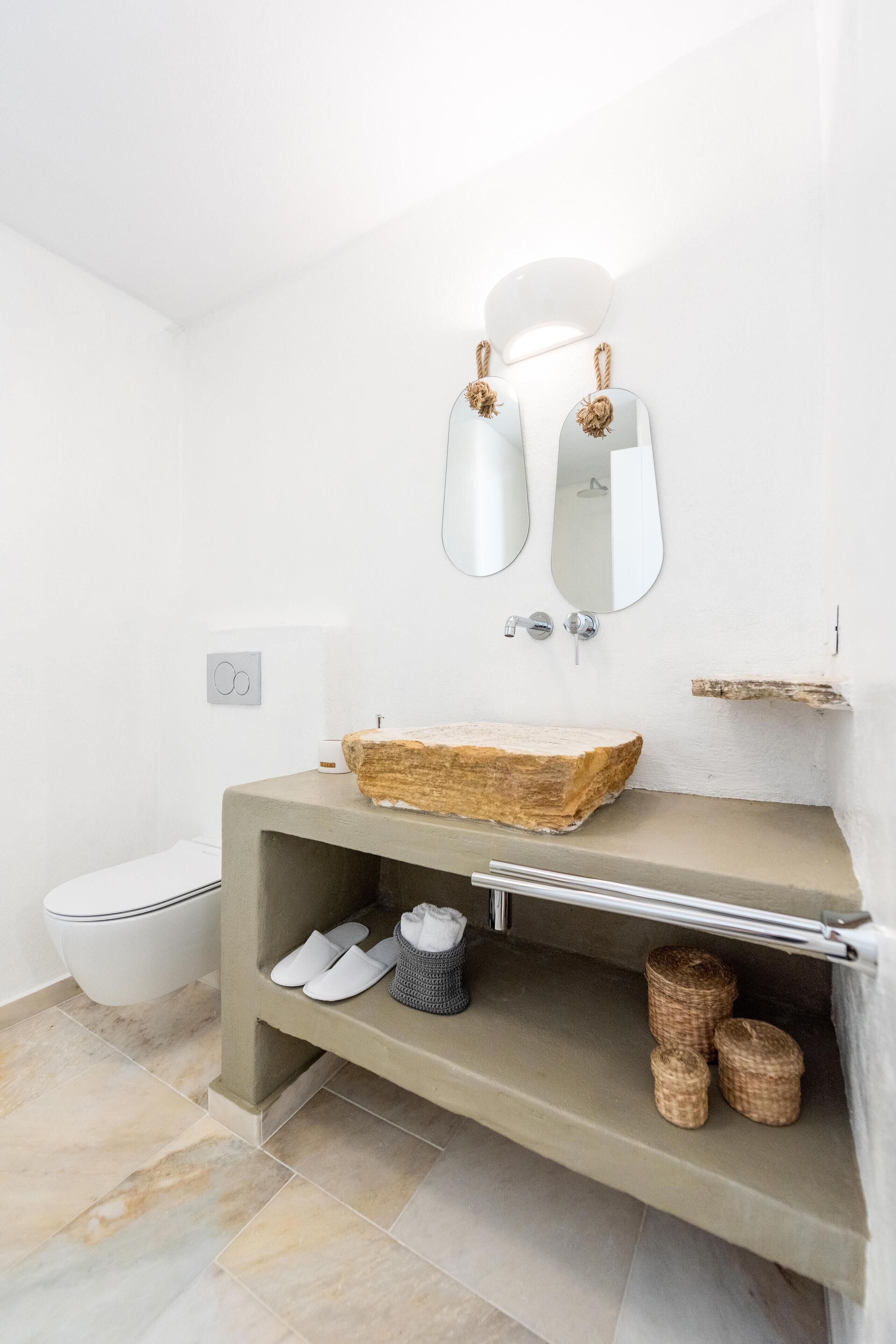 Et moderne badeværelse med en stenvask, to vægmonterede spejle, et toilet og en hylde med sko og kurve.