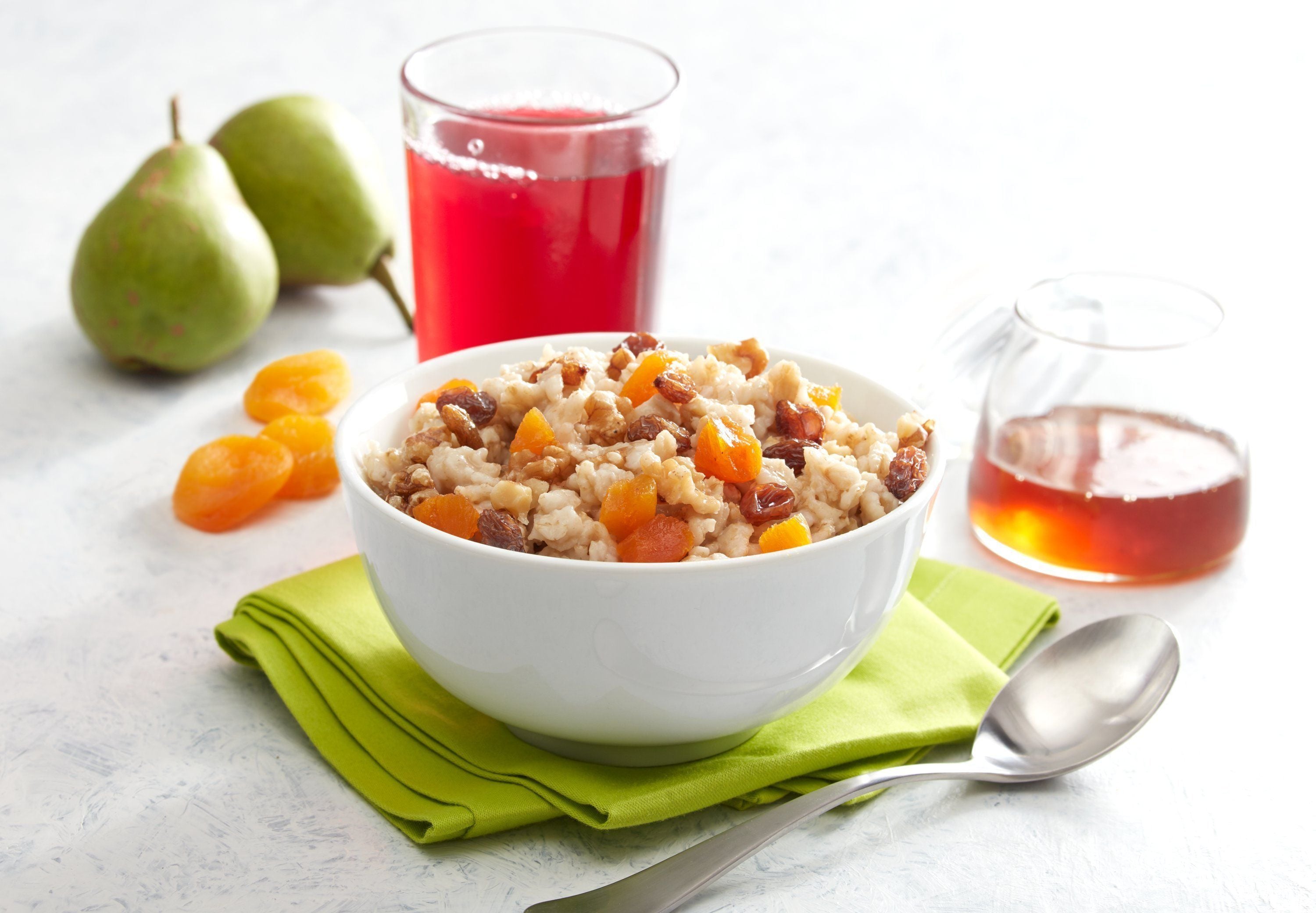 Un tazón de avena con pasas y fruta picada, un vaso de jugo rojo y un vaso de líquido transparente sobre una superficie blanca con una servilleta verde y una cuchara.