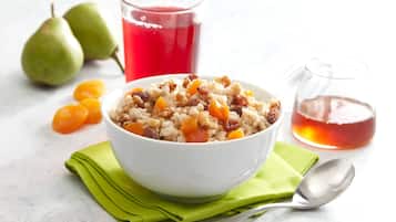 Un tazón de avena con pasas y fruta picada, un vaso de jugo rojo y un vaso de líquido transparente sobre una superficie blanca con una servilleta verde y una cuchara.