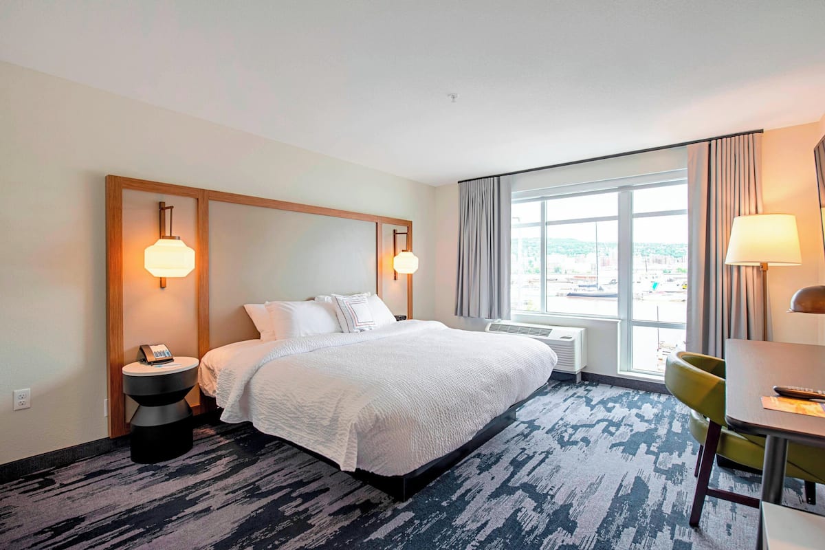 A hotel room with a large bed, bedside lamps, a desk with a chair, and a window with a view of the city.