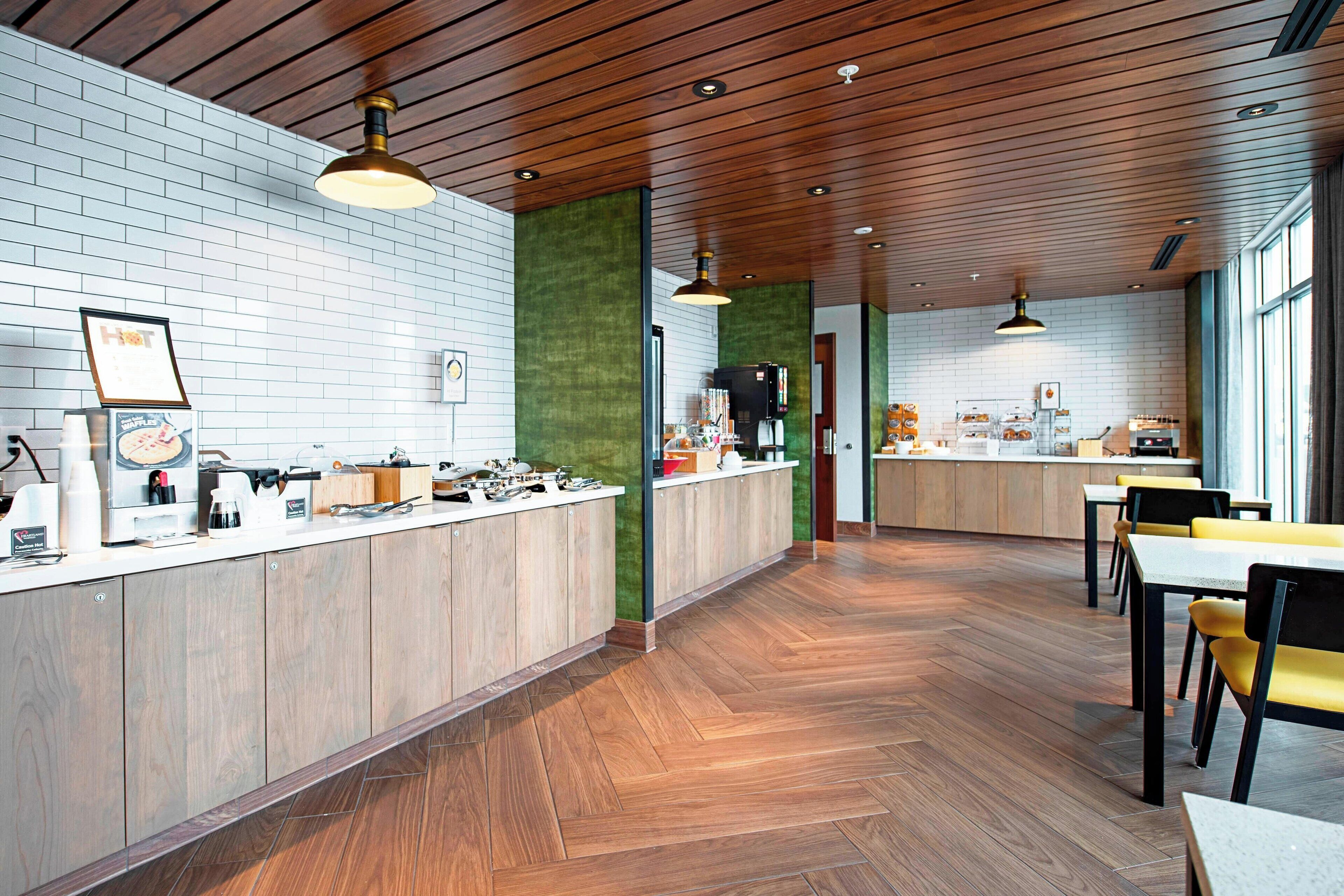 Interior moderno de restaurante com soalho de madeira, balcão com diversos alimentos e área de estar com cadeiras amarelas.