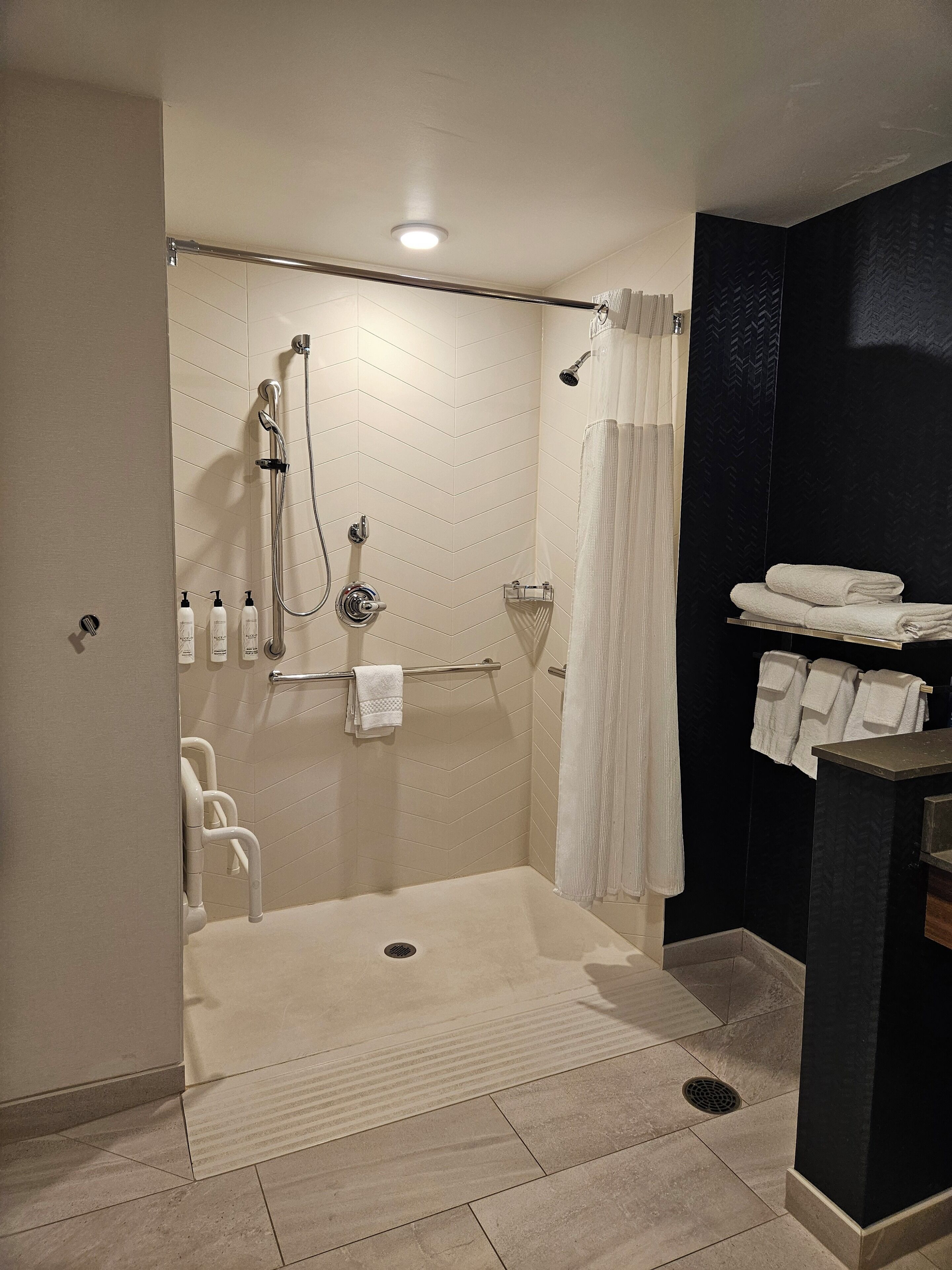 A modern bathroom with a walk-in shower, a handrail, a towel rack, and a small sink area.
