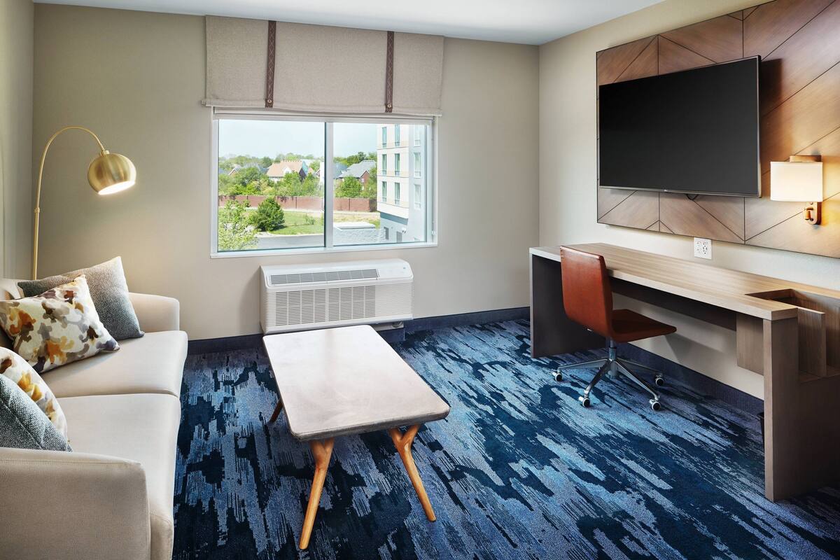 A hotel room with a sofa, a small table, a desk with a chair, a TV mounted on the wall, and a window with a view of buildings and trees.