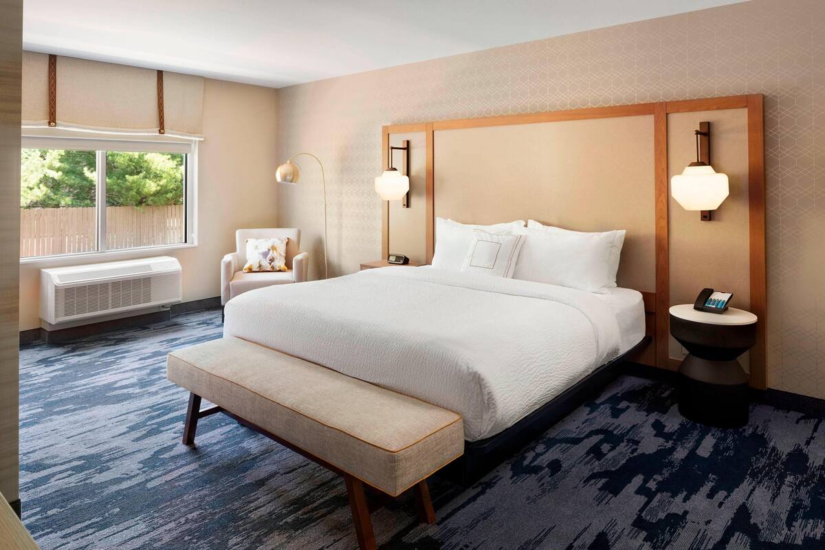 A hotel room with a large bed, a bench, a chair, a bedside table, and a window with blinds.