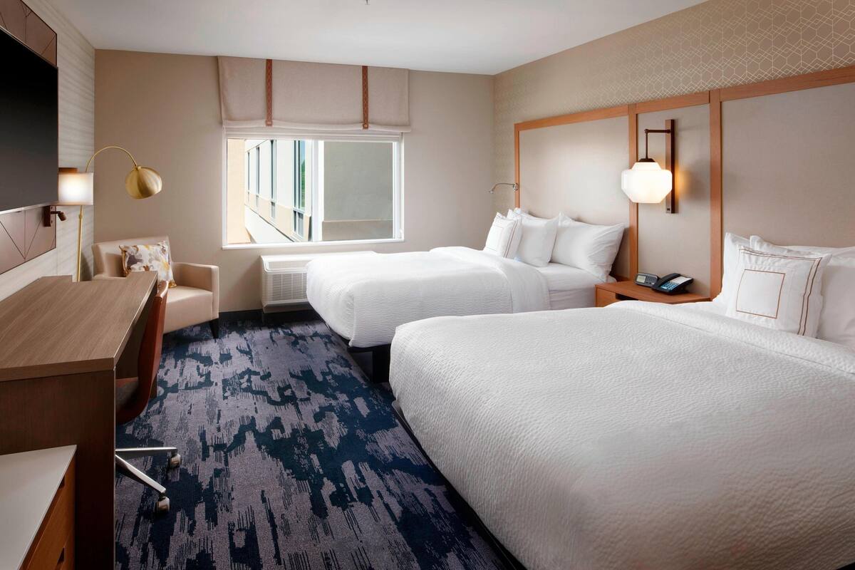 A hotel room with two beds, a desk, a chair, a TV, and a window with a view.
