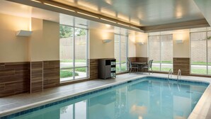 A swimming pool with clear blue water, surrounded by a tiled deck. There are chairs and a small table near the pool, and the area is well-lit with natural light from large windows.