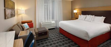 A hotel room with a large bed, a desk with a chair, a sofa, a window with curtains, and a wall-mounted lamp.