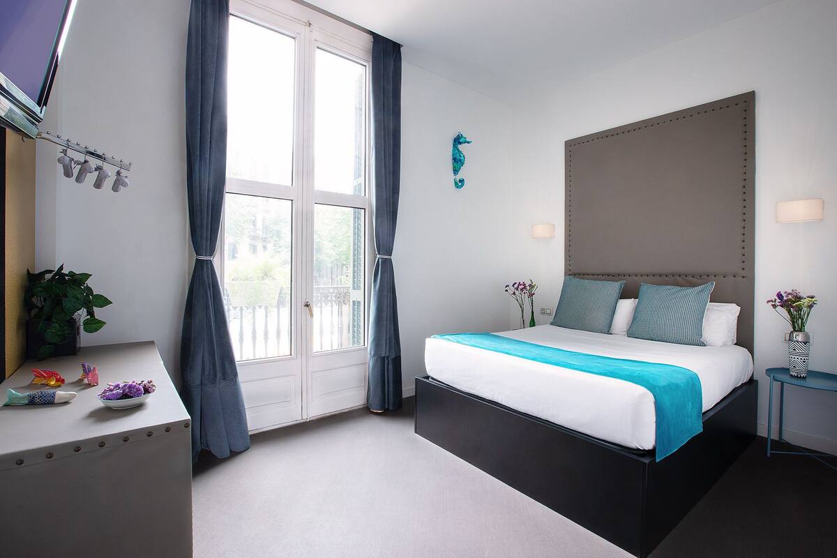 A modern hotel room with a large bed, a desk with a plant, a window with curtains, and a decorative wall piece.