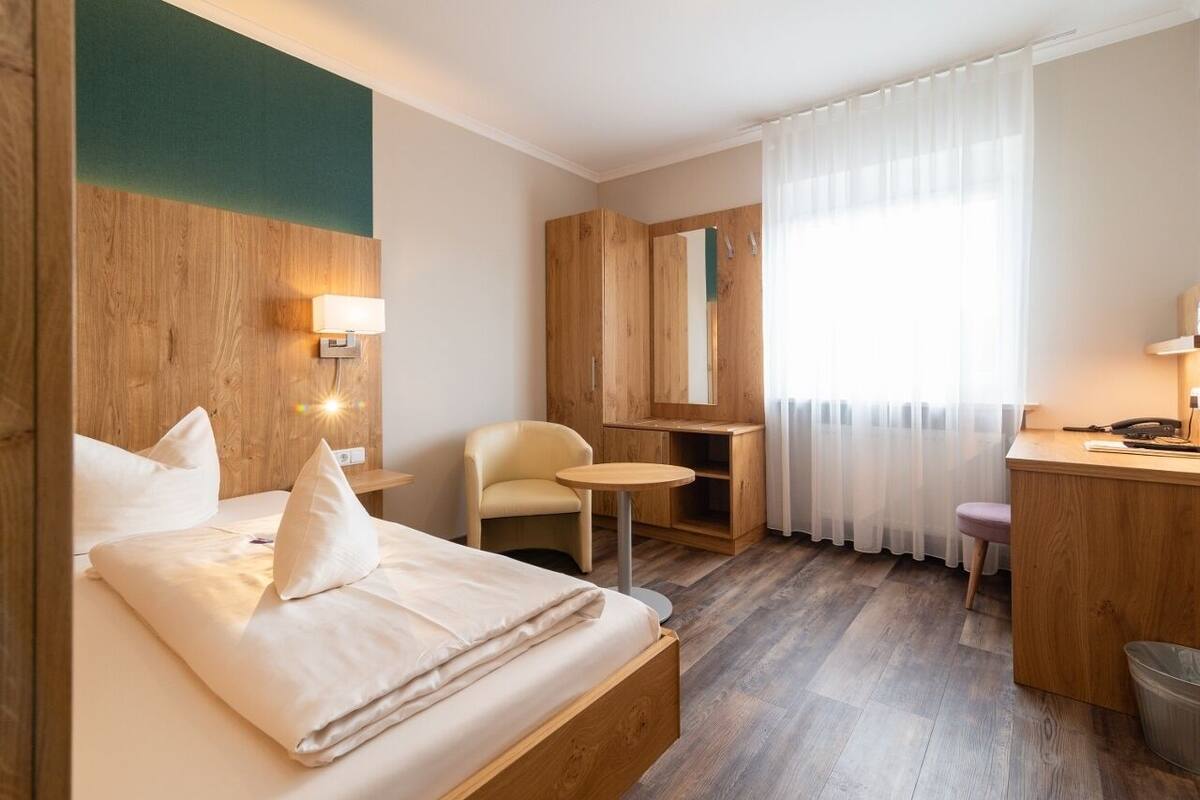 A hotel room with a wooden headboard, a bed with white linens, a small round table, a chair, a desk with a chair, a mirror, and a window with white curtains.