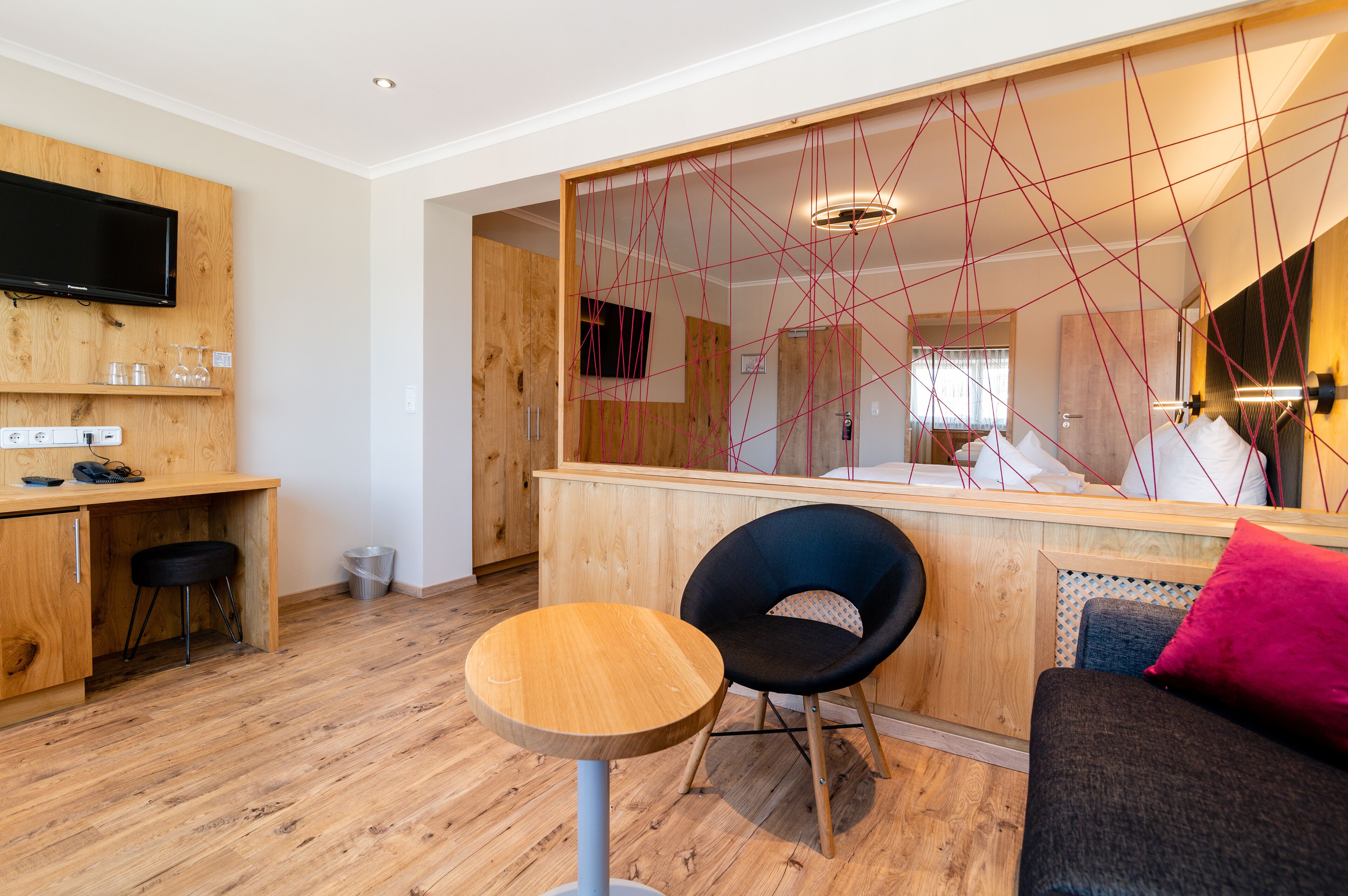 Ein modernes Zimmer mit Schreibtisch aus Holz, einem runden Tisch, einem schwarzen Stuhl und einer Couch mit einem rosafarbenen Kissen.