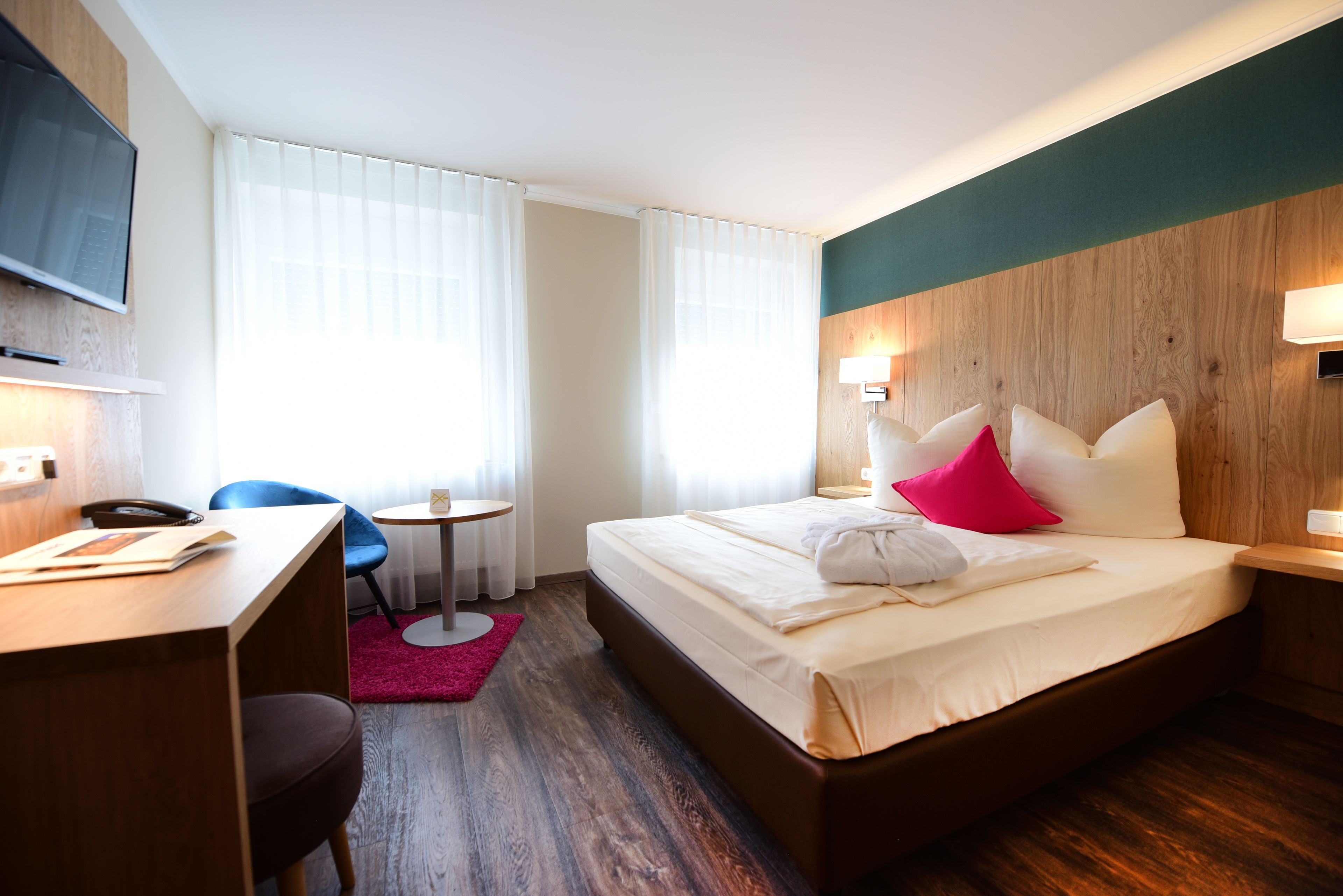 Ein Hotelzimmer mit Bett, Schreibtisch, Stuhl, Fernseher und einem Fenster mit Vorhängen.