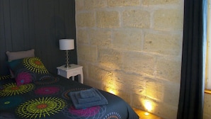Un dormitorio con pared de piedra, cama con almohadas coloridas, lámpara de noche y ventana con cortinas.
