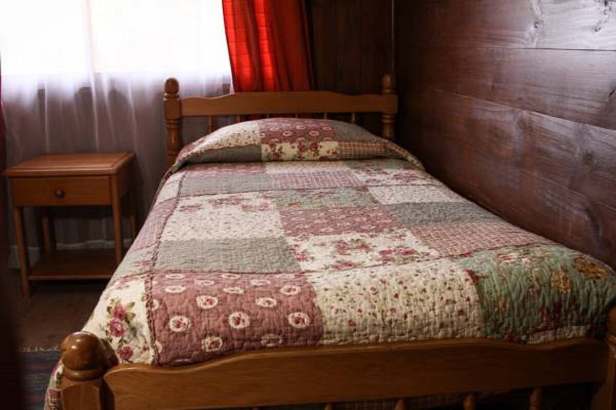 Una cama de madera con colcha, una mesita de noche y una ventana con cortinas.