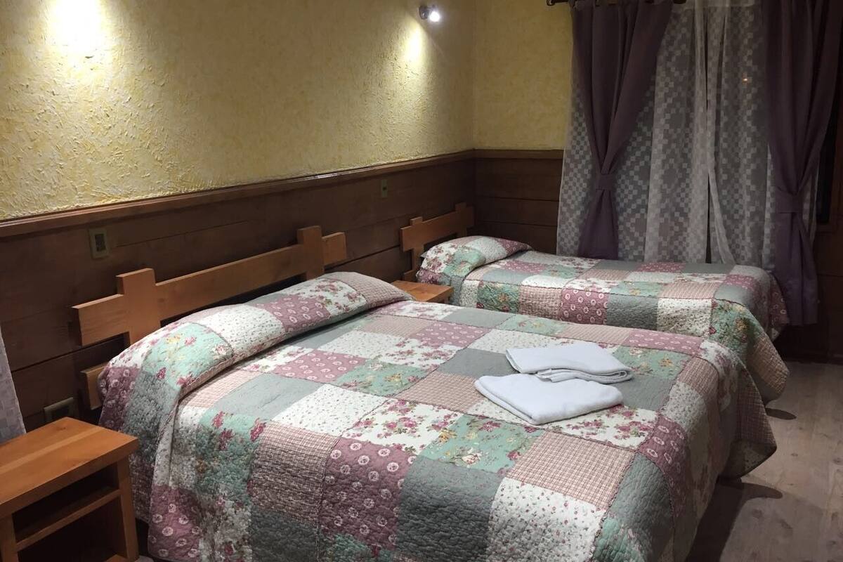 Una habitación con dos camas de madera, cada una con colchas y almohadas de estampado floral, una mesita de noche de madera y una ventana con cortinas estampadas.