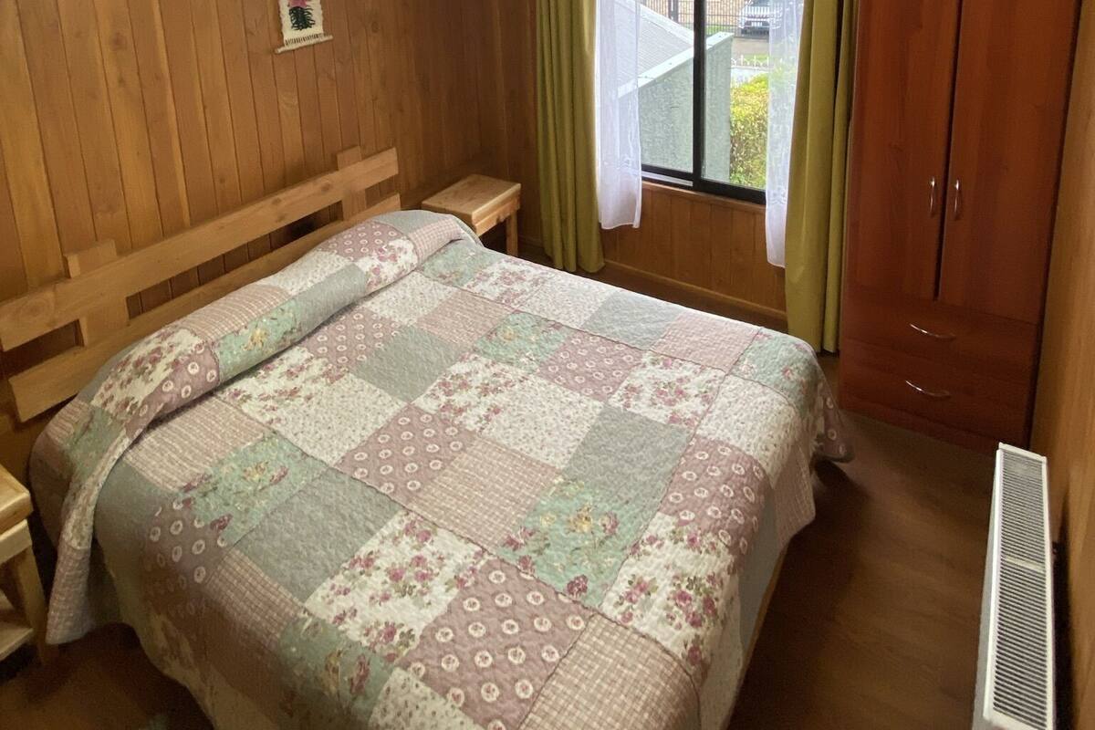 Un dormitorio con cabecera de madera, cama con colcha floral, mesita de noche, ventana con cortinas y armario.