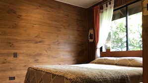 A wooden-paneled room with a bed, a window with curtains, and a wall-mounted light switch. - Hostal el Nogal Pucón (Pucón)