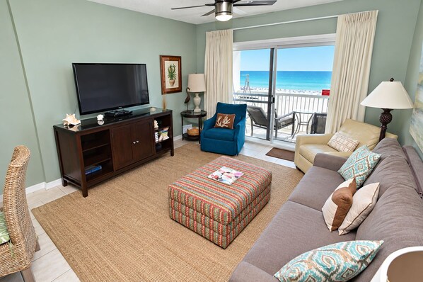 A living room with a television, sofa, ottoman, and a view of the beach through the sliding doors.