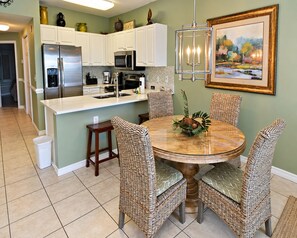 A kitchen with white cabinets, a stainless steel refrigerator, a microwave, and a coffee maker. There is a round dining table with wicker chairs and a centerpiece.
