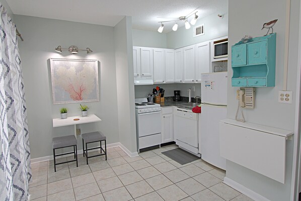 A compact kitchen with white cabinets, a stove, a microwave, and a teal-colored storage unit. There are two stools and a small table with plants. - 🌊Things are different at the beach!  Best time for a beach getaway!🌊🏖️🌴🌴 (Gulf Shores)
