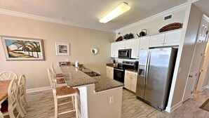 A compact kitchen with white cabinets, a stainless steel refrigerator, a microwave, and a stove. There is a small island with a sink and a dining area with chairs. - What if I told you…You CAN afford Paradise! Make the Call today! 🤙 (Orange Beach)