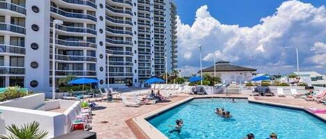 Un hotel con piscina, sillones de descanso y un edificio alto al fondo.
