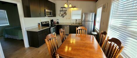 A kitchen with wooden cabinets, a dining table with chairs, a refrigerator, and a window with blinds.