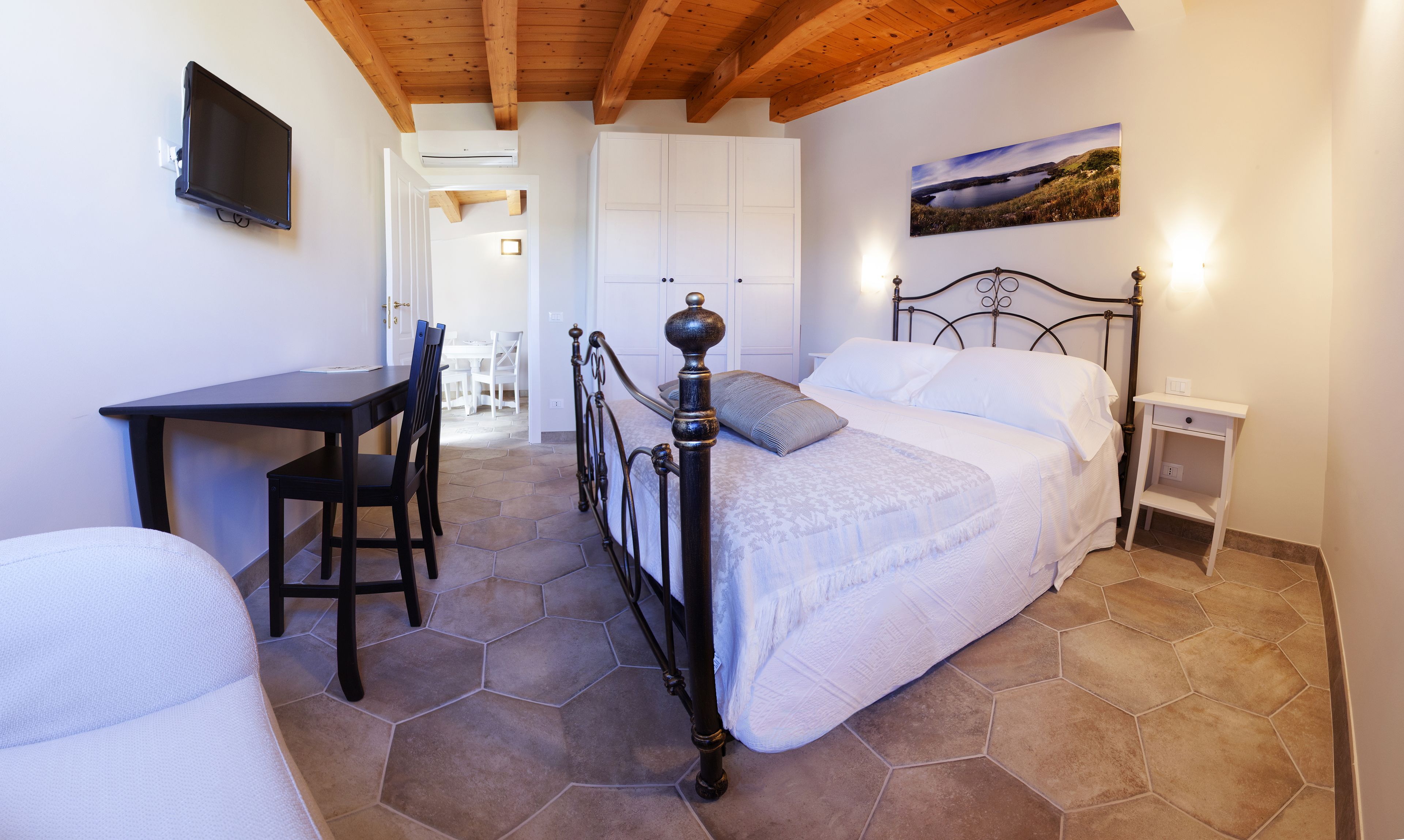 A bedroom with a bed, a desk, and chairs. There is a painting on the wall and a window with a view.