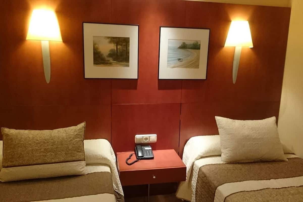 A hotel room with two beds, a bedside table with a phone, and two wall-mounted lamps.