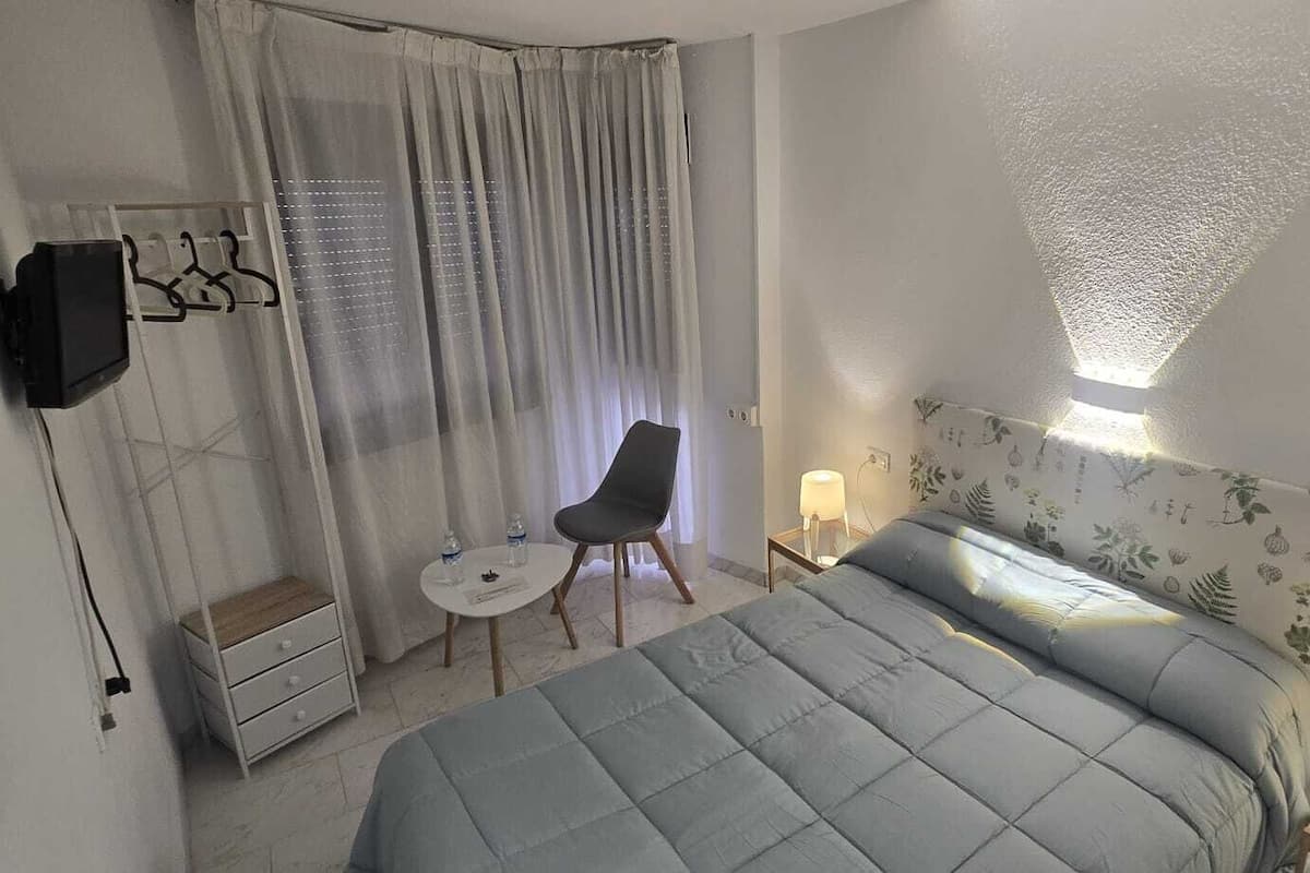 A hotel room with a bed, a chair, a small table, a TV, and a window with curtains.