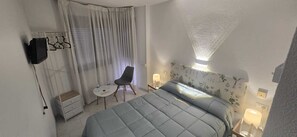 Une chambre d’hôtel avec un lit, une chaise, une petite table, un téléviseur et une fenêtre avec des rideaux.