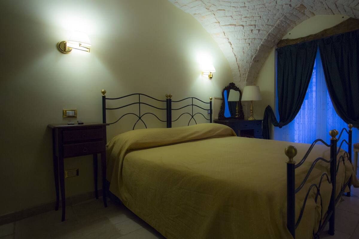 Un dormitorio con cama, mesita de noche, silla, espejo y ventana con cortinas.