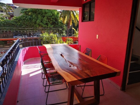 Una larga mesa de madera con sillas, frente a una pared roja, con vista a un balcón con barandilla y follaje.