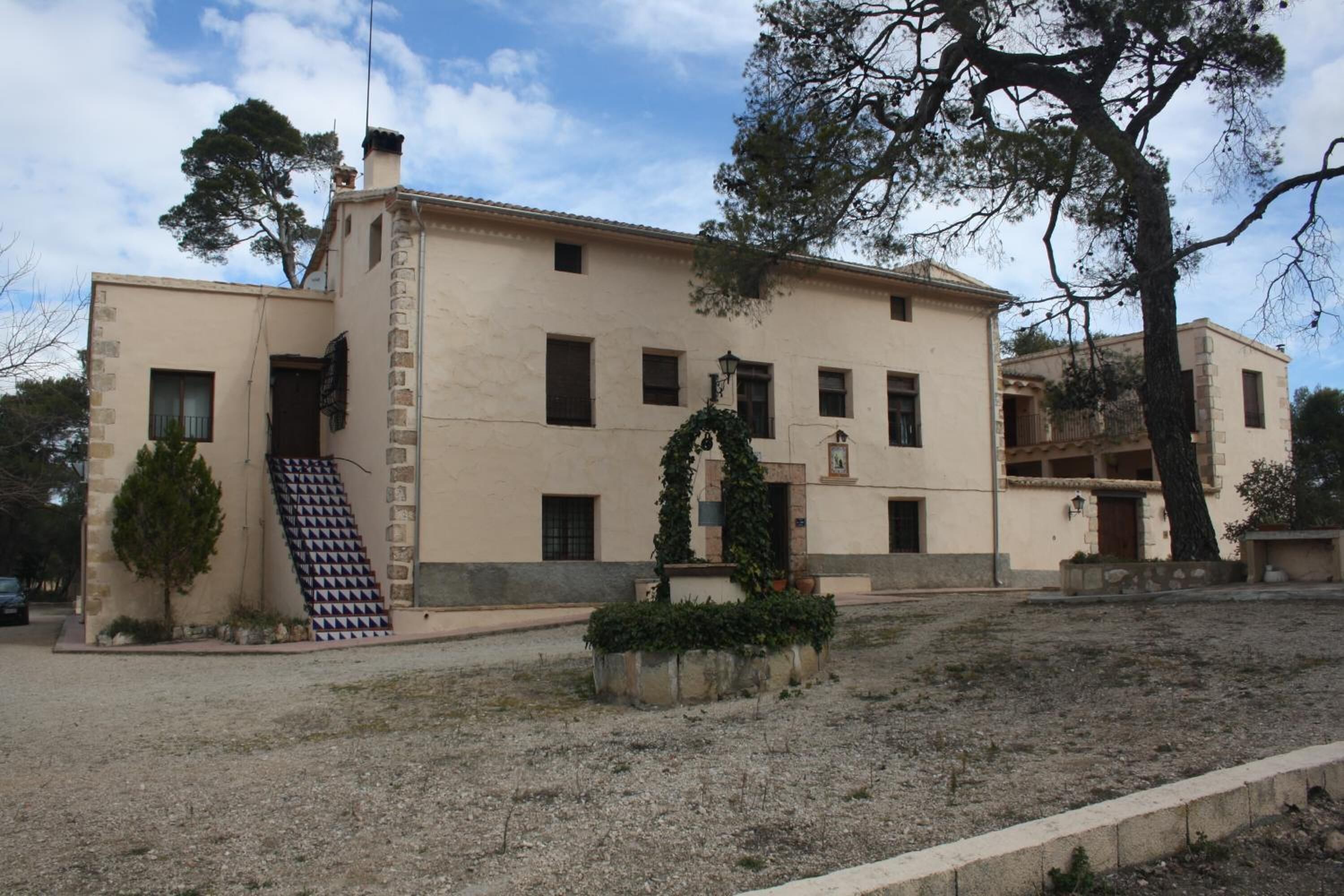 A two-story building with a beige facade, a staircase with patterned tiles, and a small garden area with a tree and shrubs.