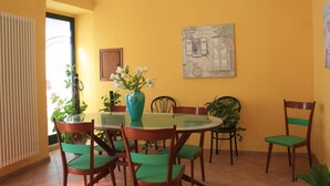 Een eetgedeelte met een ronde tafel, groene fauteuils, een blauwe vaas met bloemen, en een schilderij aan de muur.