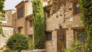 Un edificio de piedra con un patio, destacando un árbol alto, una puerta de madera y una ventana pequeña.