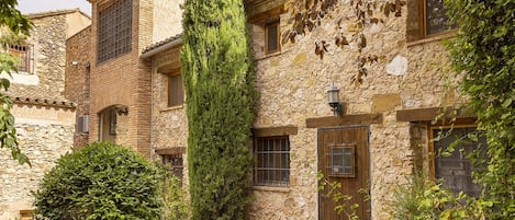 A stone building with a courtyard, featuring a tall tree, a wooden door, and a small window.
