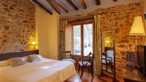 Un dormitorio con pared de piedra, vigas de madera, una cama con almohadas, una silla, un televisor y una ventana con cortinas.
