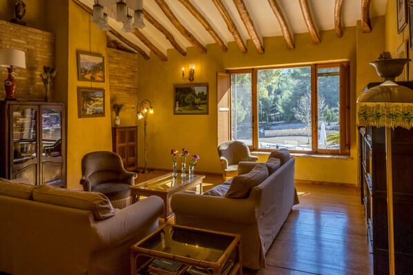 A cozy living room with a sofa, armchairs, a coffee table, and a fireplace. There are framed pictures on the walls and a window with a view of trees. - Hotel Rural Mas Fontanelles (Biar)