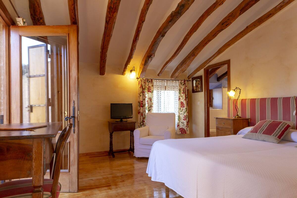 A cozy bedroom with a wooden ceiling, a bed with striped pillows, a bedside table with a lamp, and a television on a small table.