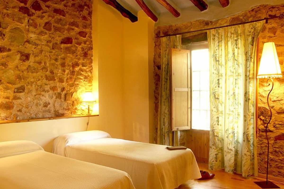 A room with two beds, a stone wall, a wooden floor, and a window with curtains.