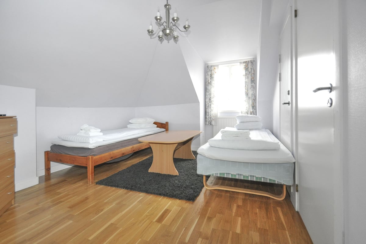 Una habitación pequeña y luminosa con piso de madera, una cama, una mesita y una ventana con cortinas.