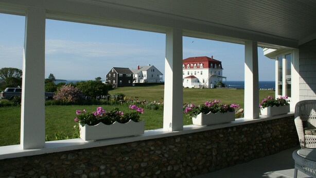 A covered porch with stone walls and white columns, overlooking a grassy area with a large white building and a red-roofed structure in the distance. - Rose Farm Inn (Block Island)